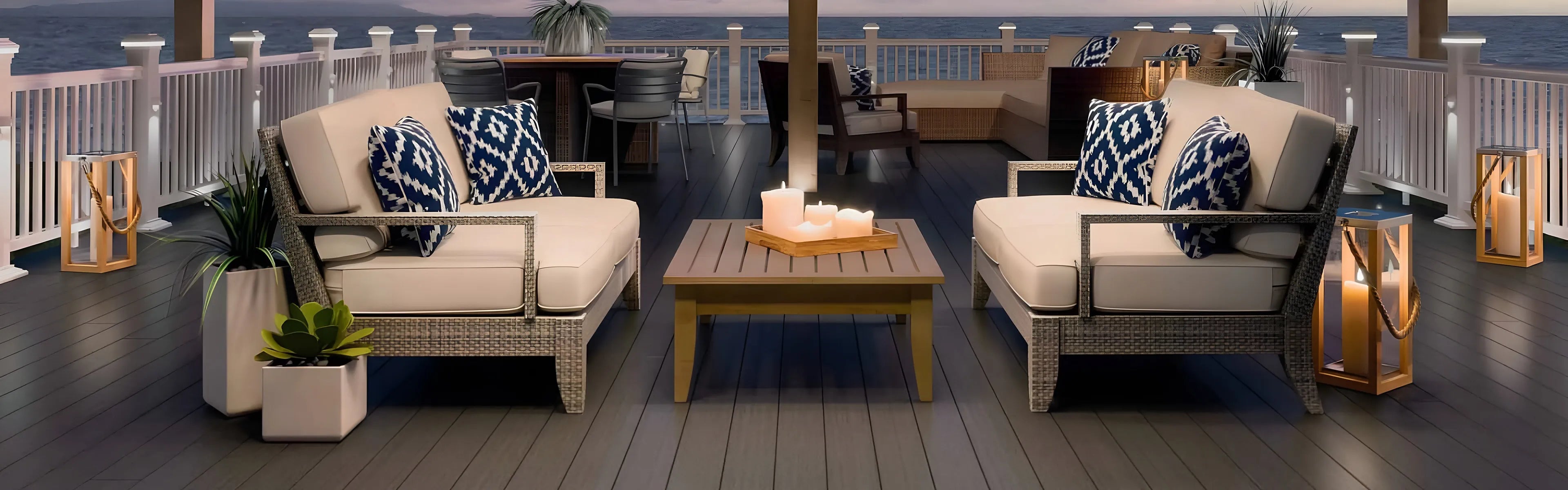 Outdoor patio with cushioned chairs, patterned pillows, a wooden table with candles, and ocean view at sunset. Lanterns, plants, and more seating are in the background.