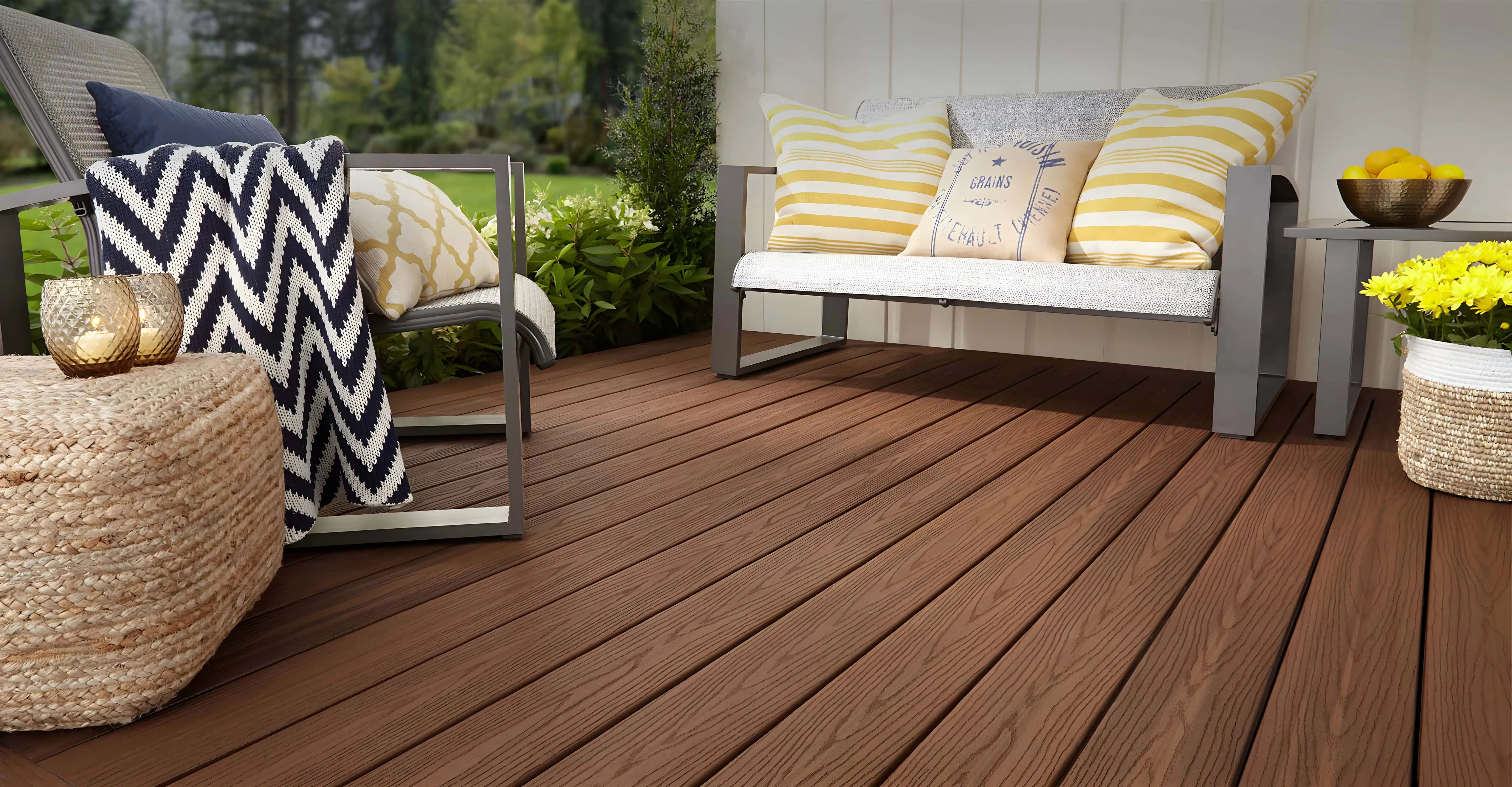 Outdoor patio with brown wood alternative decking, metal-framed seating, cushions, a throw blanket, and potted yellow flowers.