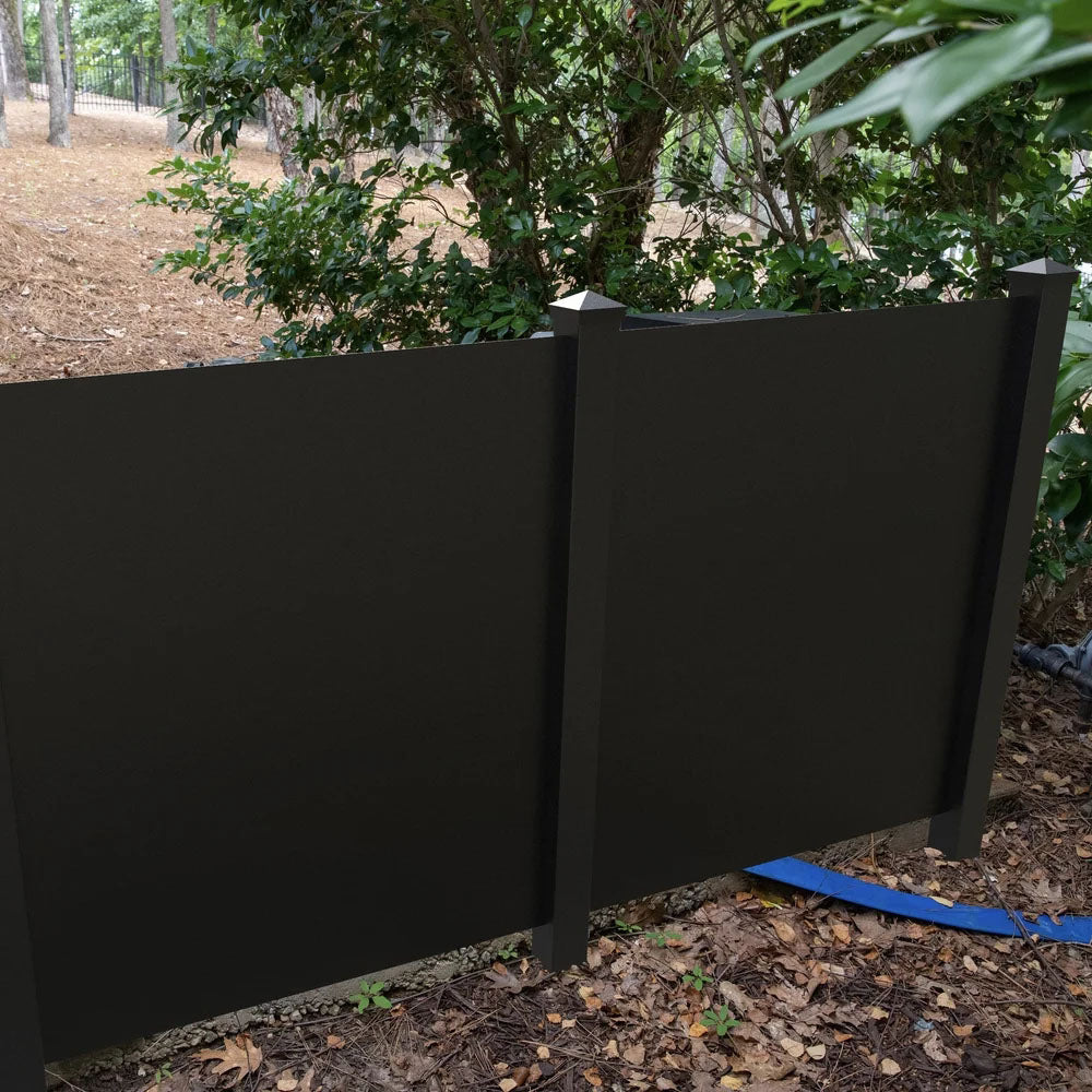 A Sunbelly Solid Shorty privacy screen stands among green bushes and trees with dry leaves scattered on the ground.