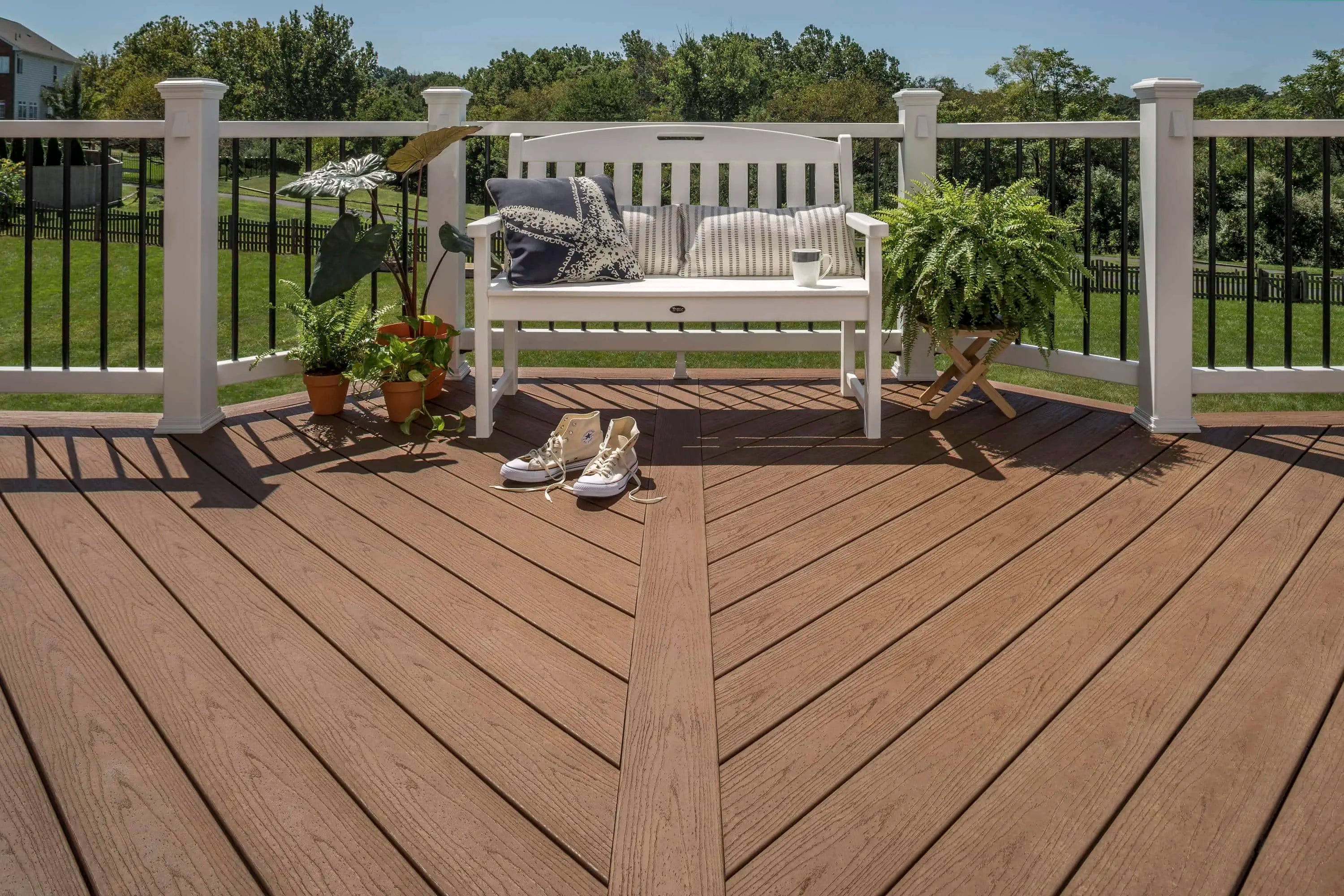 A white bench with cushions and plants sits on a brown composite deck with white railings overlooking a grassy yard.